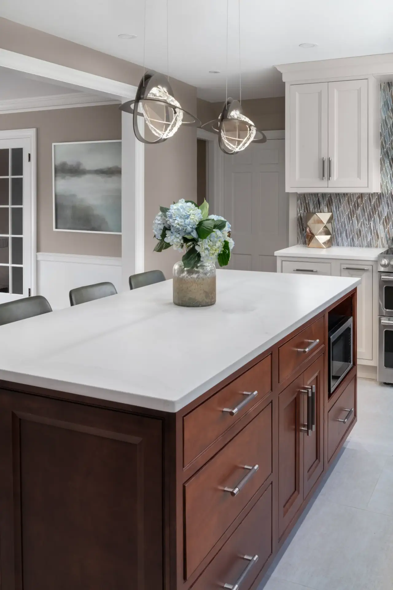 A spacious kitchen featuring a dark wood island with white countertop, pendant lighting, blue hydrangea flowers in a vase, and white cabinetry in the background.