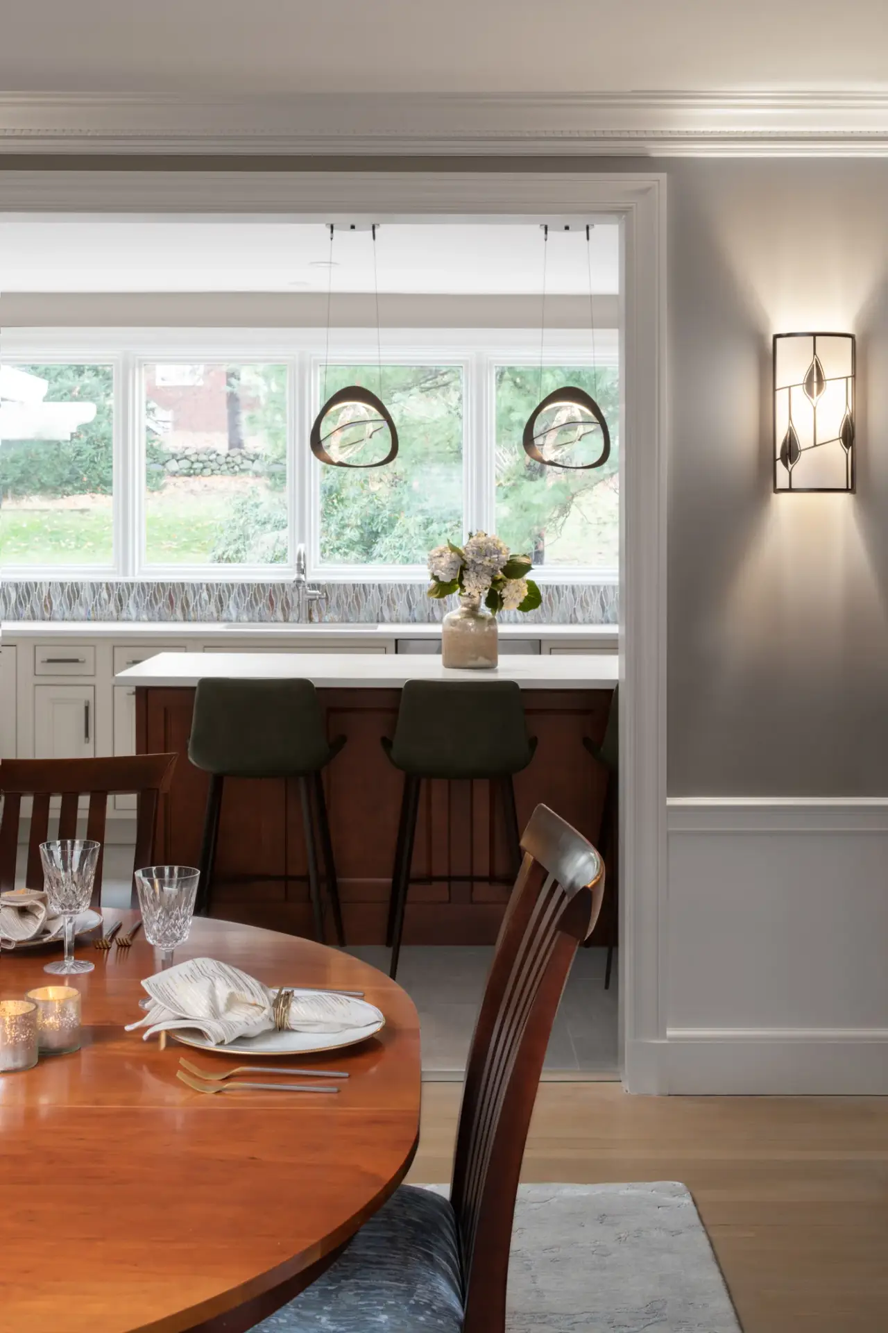 Contemporary kitchen viewed from dining room with large windows, white cabinetry, dark bar stools at island, pendant lights, and garden views.