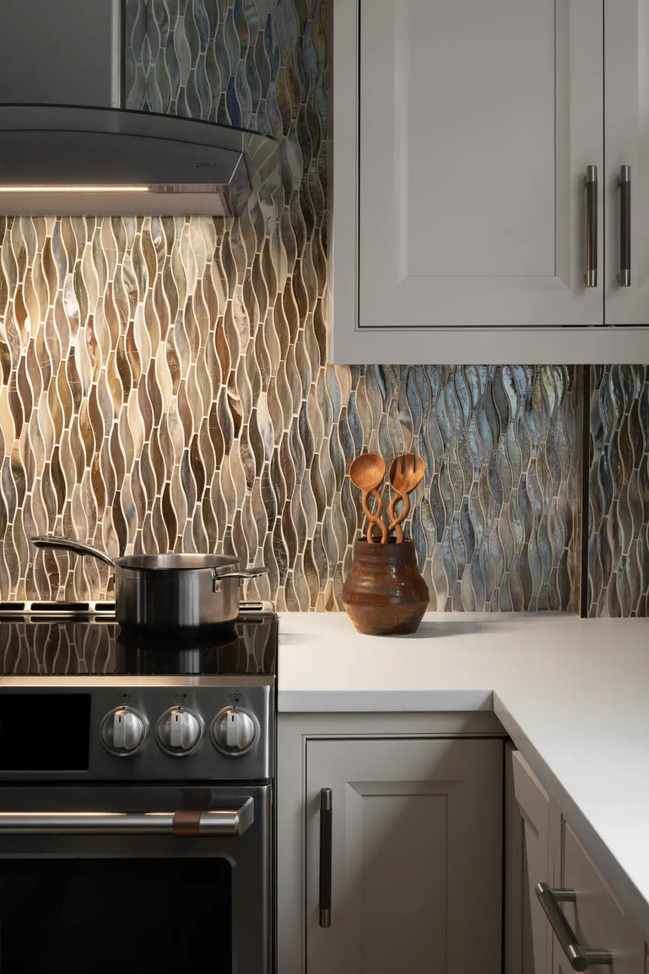 Contemporary kitchen featuring wavy patterned backsplash tiles, black range with pot, white countertop, gray cabinetry, and wooden utensil holder.