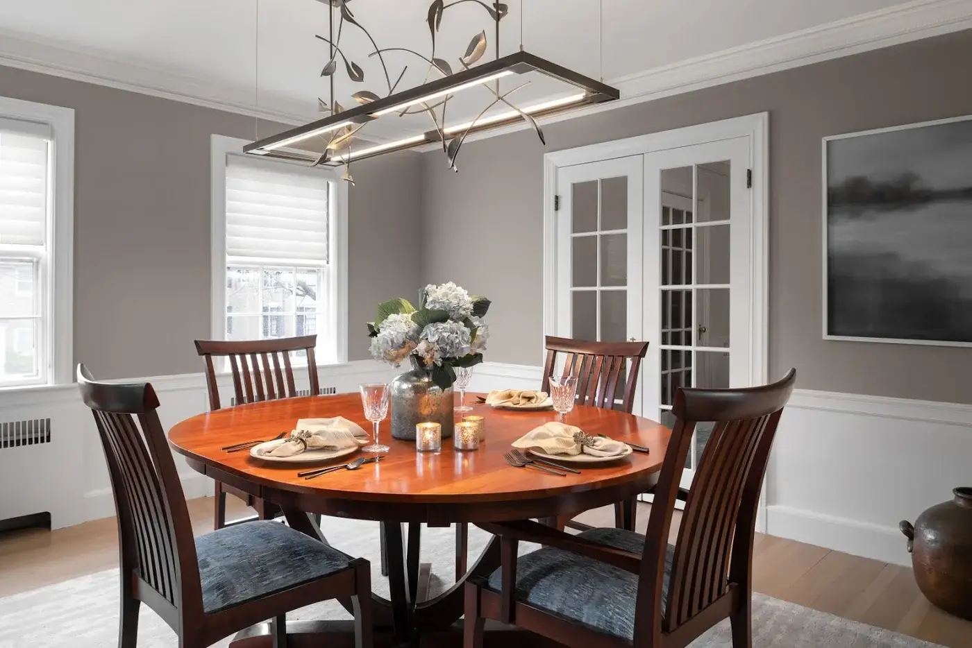 A modern dining room features a polished round wood table set for four, with matching chairs, white hydrangeas in a vase, and an artistic pendant light above.