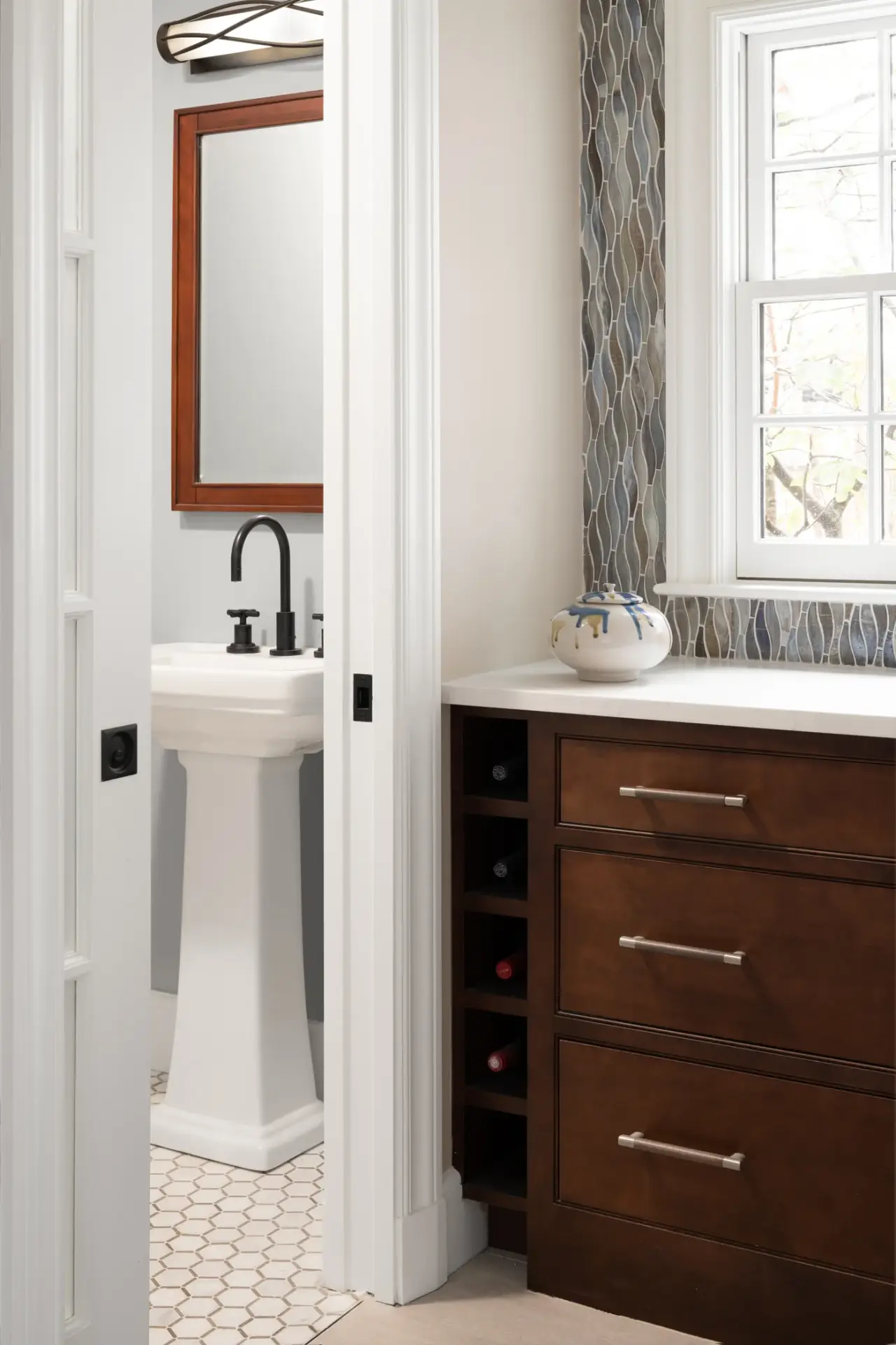 A modern bathroom features a pedestal sink with black faucet and wooden-framed mirror on the left, and a dark wood vanity with drawers on the right, separated by a patterned gray curtain panel and window.