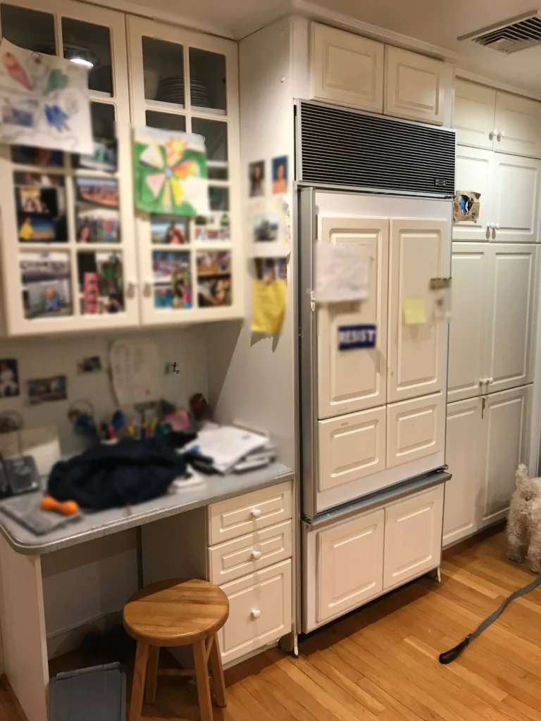Kitchen interior featuring cream-colored cabinetry, stainless steel refrigerator with photos and notes on door, built-in desk workspace with storage, wooden bar stool, and hardwood flooring.