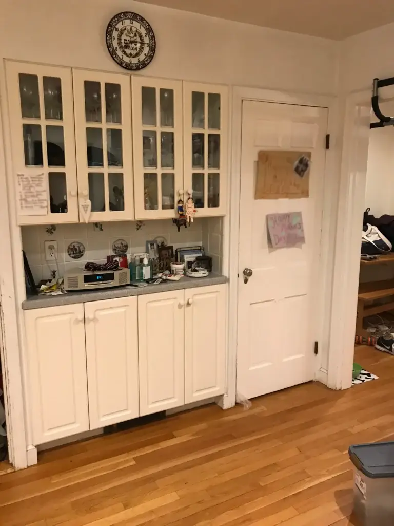A white painted kitchen hutch with glass-fronted upper cabinets and solid lower cabinets featuring a gray countertop, decorated with dishes, photos, and collectibles, against a beige wall.