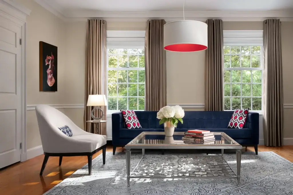 Bright living room featuring a navy sofa with red and white patterned pillows, white accent chair, glass coffee table, white pendant light with red interior, and large windows with taupe curtains.