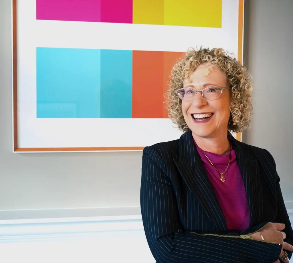 Woman with curly blonde hair wearing glasses, black pinstriped blazer, and purple top smiling in front of abstract colorful geometric painting.